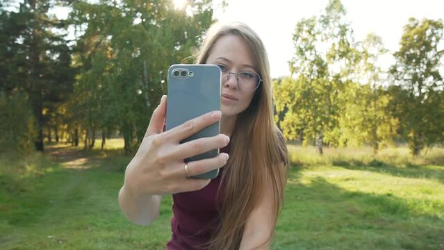 Blonde Girl blogger with glasses making selfie on a smartphone, outdoors in a forest park. Online, social lifestyle. Zoom in rack focus shot. 