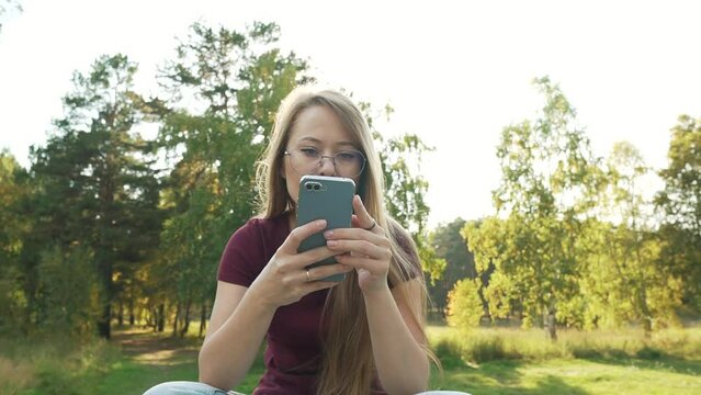 Relaxed woman using smartphone in a park, reading online news or social media. Girl chatting in mobile phone messengers outside, sitting in a park. Zoom out, rack focus shot.