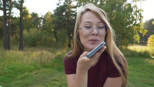 Smiling woman using smartphone and laughing, recording audio message media for social messenger. Girl sharing news mobile phone, talking in a park outside. Pan shot.  