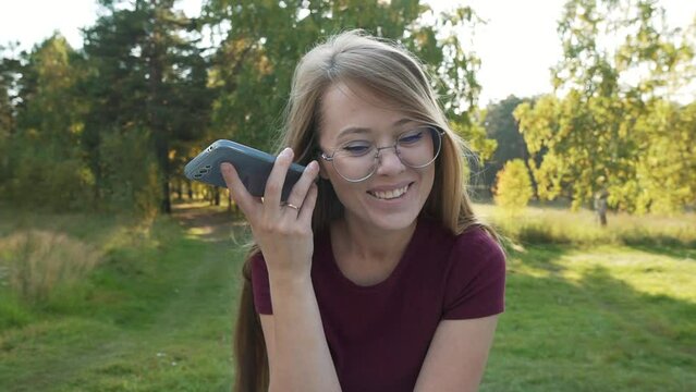 Smiling woman using smartphone and laughing, listening audio message media from social messenger. Girl holding mobile phone sitting in a park outside. Pan shot. 