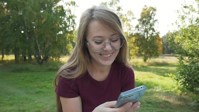 Smiling woman using smartphone and laughing, listening and answering audio message media from social messenger. Girl holding mobile phone sitting in a park outside. Pan rotation shot.   