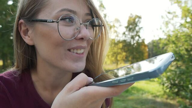 Smiling woman having fun and laughing, answering and recording audio message media from social messenger. Girl using smartphone sitting in a park outside. Zoom out.