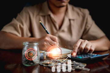 Man using calculator to calculate and money stack, coin in a jar, lightbulb on desk with note book. idea saving energy and accounting finance concept.