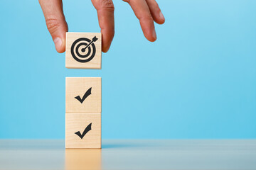 Hand putting stack of wooden blocks with marks and target icons on a blue background copy space. Business development strategy, advancement and goal concept.