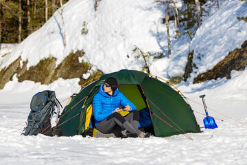 A man with a smartphone in a green tent with a backpack and a snow shovel in the spring forest. A...