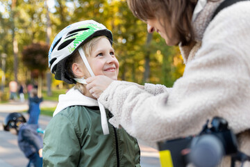 Close-up mom parent hand help put on and fasten safety helmet on cute blond caucasian girl riding bike or scooter city street park outdoors on autumn fall day. Child sport activity protection care