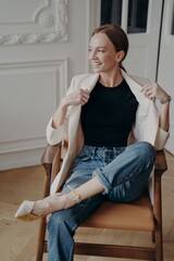 Successful woman is sitting in armchair with her legs crossed. Stylish young adult european lady.