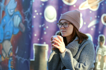 A girl in warm clothes and a hat, glasses, drinks from a cup of coffee in an autumn park
