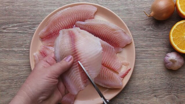 Pink tilapia fish, large pieces of fish fillet, close, lies on a plate, the cook's hand cuts the bones out of the fish with scissors.