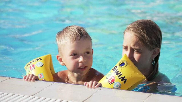 Close up of happy children swimming in pool. Brother and sister. Young beautiful girl and little boy laugh and smile broadly. Boy in inflatable armbands is holding onto side of pool. Look into camera.