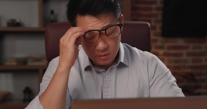 Tired Asian man takes off glasses, uses a laptop and having a work problem