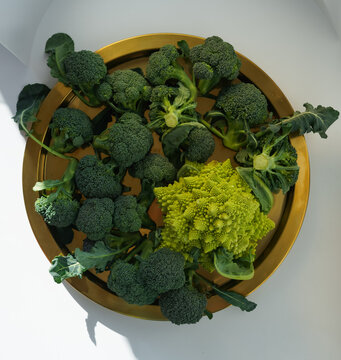 golden round tray with harvest of green fresh broccoli with leaves and romanesco in daylight on light background