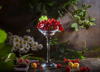 Red currants in a glass wine glass, raspberries and a bouquet of daisies on a wooden table