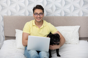 Smiling happy man sitting on the bed and hugging his lovely pet black pug breed and working on laptop. Friendship concept