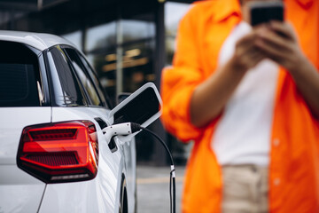 Woman charging electric car at the charging station and using mobile phone