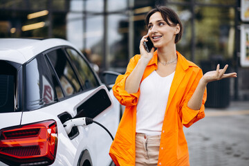 Woman charging electric car at the charging station and using mobile phone