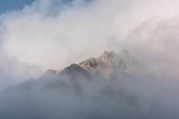 Lovely scenery with big mountain peak in thick clouds in sunlight. Sunlit high pinnacle in gantly cloudy sky. Large mountain top in clearance of dense fog. Beautiful rocks in low clouds in sunrise.