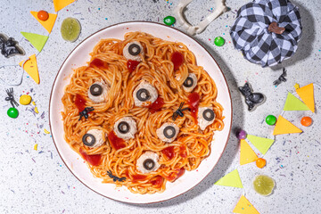Creepy funny pasta spaghetti for Halloween party dinner. Spaghetti with tomato sauce and chicken meatball decorated with black olives like eyes, white table background with Halloween decorations