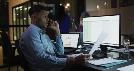 Video of biracial businessman talking on smartphone and using computer at night in office - Powered by Adobe