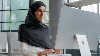 Young arab businesswoman in hijab working on computer smiling enjoying office work successful woman professional manager company employee develops project online searching for information on Internet