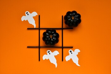 Creative tic tac toe game made of halloween decorations. Ghosts and  black pumpkins on orange background. Autumn party concept, children game. Top view, flat lay - obrazy, fototapety, plakaty