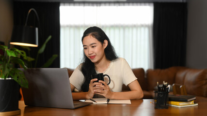 Asian female freelancer holding cup of coffee and reading email on her laptop