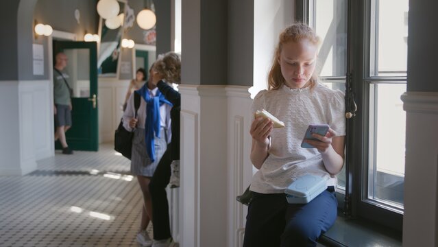 Teenage Girl Having Lunch And Surfing Internet On Smartphone Sitting On Windowsill At School