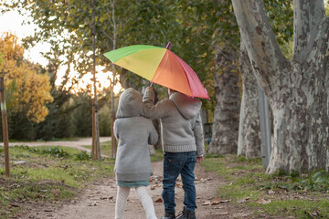 dos gemelos paseando por un parque en otoño con un paraguas aco iris	