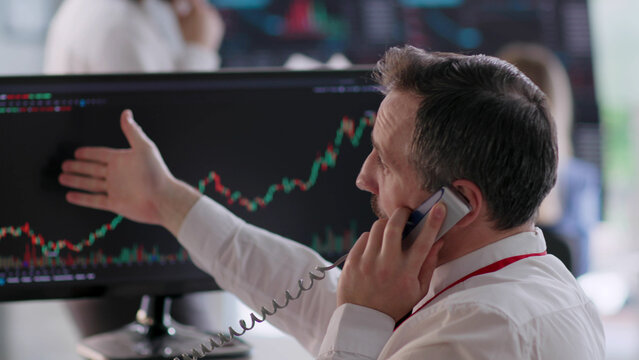 Back view of stock broker looking at computer monitor with graphs and talking on phone