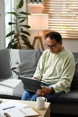 Happy mature man sitting on couch and checking email, reading online news on tablet. Elderly technology concept