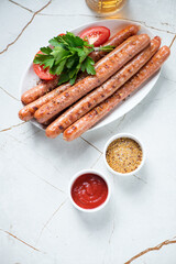 Barbecued sausages served with fresh parsley, tomatoes, dips and beer on a light-beige stone background, high angle view, vertical shot