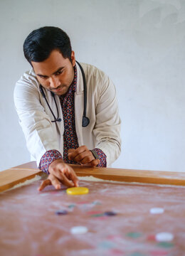 South asian young doctor passing his leisure time by playing carom board, Bangladeshi medical student wearing apron and stethoscope playing indoor game