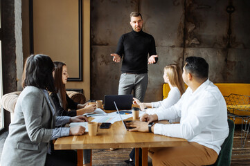 Teamwork. A group of young people are sitting at a table in a modern bright open space office. Young employee is explaining something while standing in front of an audience