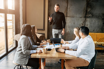 Young successful team leader gives a speech to employees. He is standing in front of a table in a modern office while the rest are sitting.