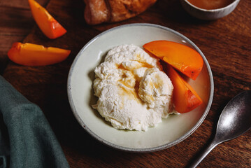 Homemade ricotta cheese or cheese curd served with fresh persimmon and honey and croissante on wooden background. Healthy breakfast. Haze effect. Selective focus