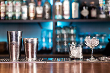 A glass of ice and a shaker stand on the bar counter. Glasses are cooled to serve a cocktail