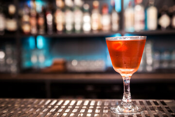 An alcoholic cocktail of red color with a decoration of lemon peel stands on a bar counter.