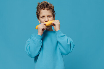 a cute, funny boy of school age stands on a blue background in a blue sweater and tries to open a banana. Horizontal studio photography with blank space for advertising mockup insert