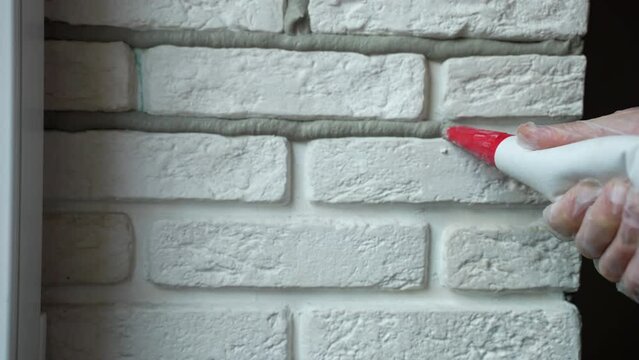 Repairing process of interior wall with white decorative stones, builder applying grout between joints, using construction syringe. Filling joints with mortar for further grouting, home interior