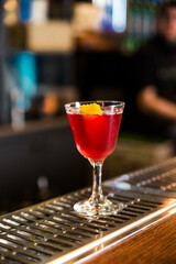 An alcoholic cocktail of red color with a decoration of lemon peel stands on a bar counter