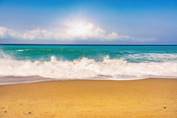 Beautiful summer background image of the Mediterranean coast with spectacular waves, golden sandy beach and bright sun on blue sky with clouds.
