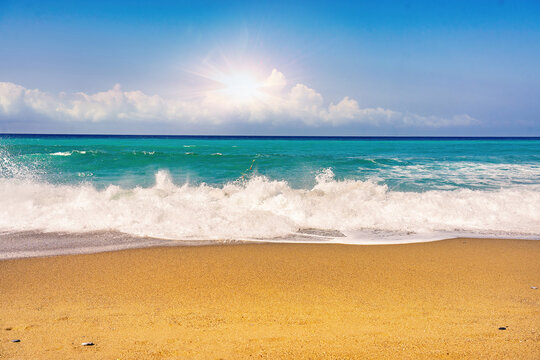 Beautiful summer background image of the Mediterranean coast with spectacular waves, golden sandy beach and bright sun on blue sky with clouds.
