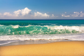 Beautiful seascape with with golden sandy beach, blue sky with clouds, horizon line and waves of surf with caps of white foam.