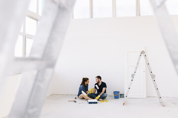Young couple in love, the newlyweds are making repairs in an empty room, painting white walls,...