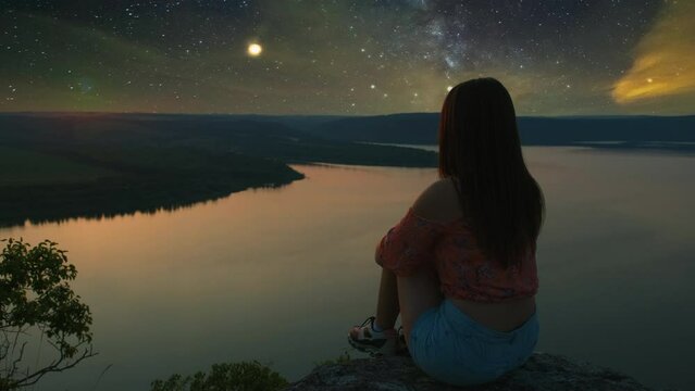 The girl sitting on the mountain top above scenic river on starry sky background
