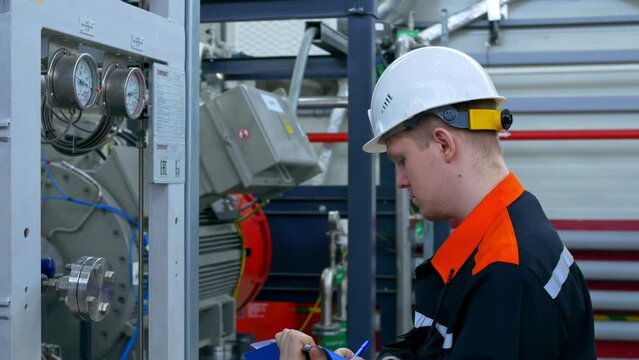 An engineer in a white helmet and overalls holds records of data on the operation and parameters of equipment at the plant. The engineer makes entries in the journal of industrial equipment.