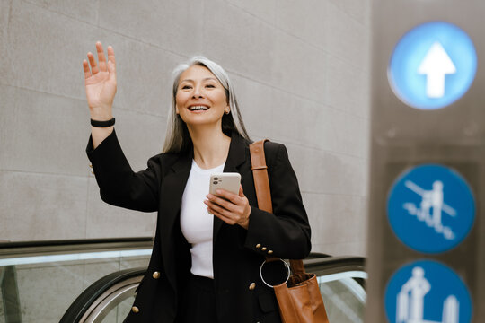 Mature asian woman gesturing and using cellphone on escalator