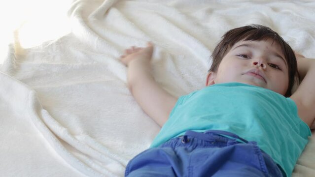 kid lies on bed morning sunshine day.child moving hands and legs similar of doing snow angel.little boy feeling safe secure cozy comfort at home with family mother.ivory blanket blue clothes