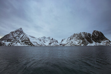 Fototapeta na wymiar Famous mountain of Lofoten covered by snow in winter season. The big rock has piramidal form and it is near the arctic sea in Norway. Small fishing boat passing by the sea in cod fishing season