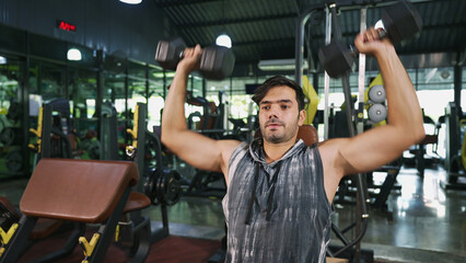 Portrait of Asian male bodybuilder sitting on fitness bench and doing dumbbell shoulder press exercise. Shot of athlete man focus on lifting double dumbbell to improve strength of his shoulder muscle.